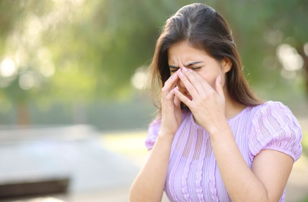 A person rubbing her eyes and scrunching her nose with green trees in the background